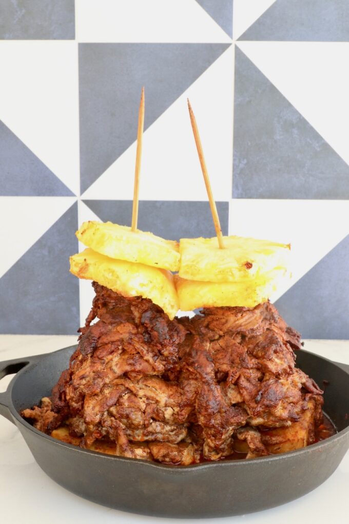 A stack of marinated, cooked pork layered on skewers, topped with pineapple slices, in a cast iron skillet.