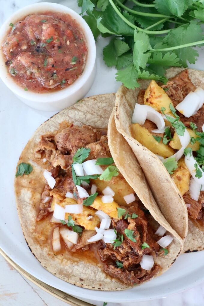 Two tacos with pork al pastor, pineapple, onion, and cilantro on tortillas, accompanied by a bowl of salsa and fresh cilantro, arranged on a plate.